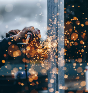 A person is using a welding tool on a piece of metal. Bright sparks are flying in all directions, creating a bokeh effect against a blurred background. The image captures the intensity and focus needed for welding work.