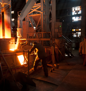 A worker in protective gear operates machinery at a foundry. Bright molten metal pours from a spout into a container, illuminating the dim, industrial workshop. Another person stands in the background observing the process.