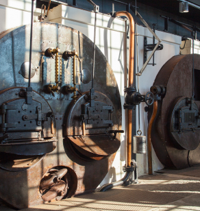 Industrial machinery with two large, rusted metal circular components and various pipes and valves. The setting appears to be an old factory or mechanical workshop with sunlight streaming in through windows, casting shadows on the equipment.