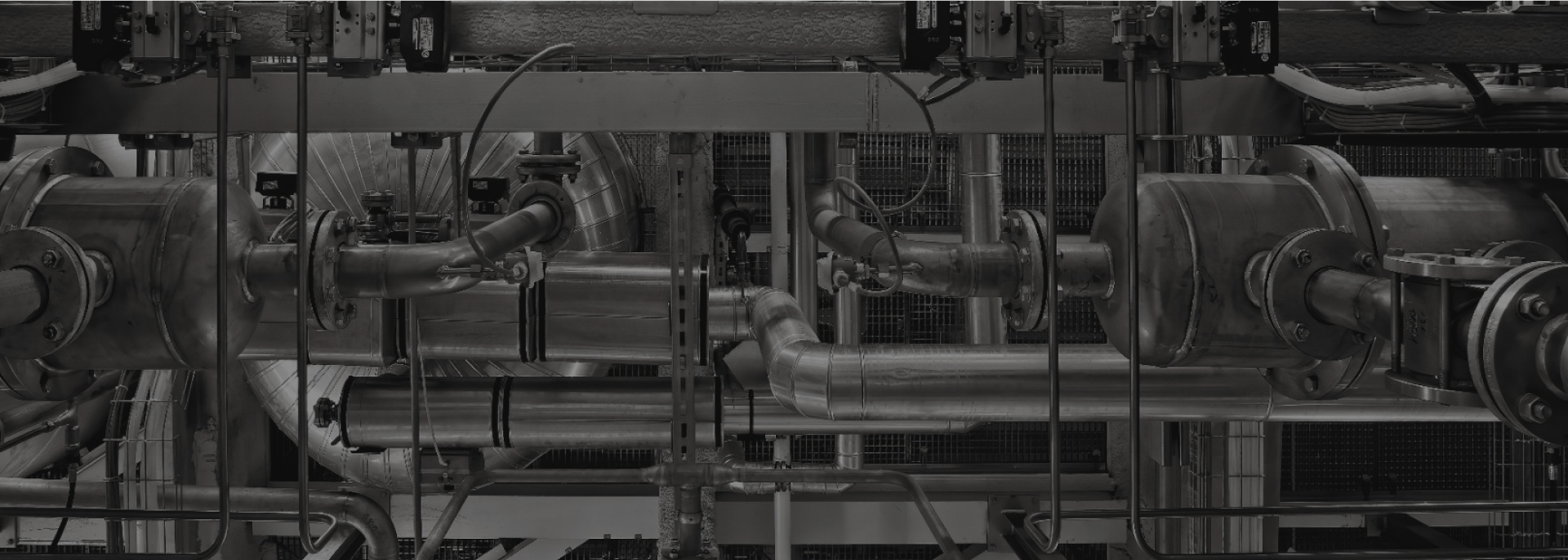 A complex network of industrial metal pipes and machinery, including valves and fittings, forms an intricate system. The pipes intersect and connect at various points, and wires are visible hanging throughout the structure. The image is monochromatic.