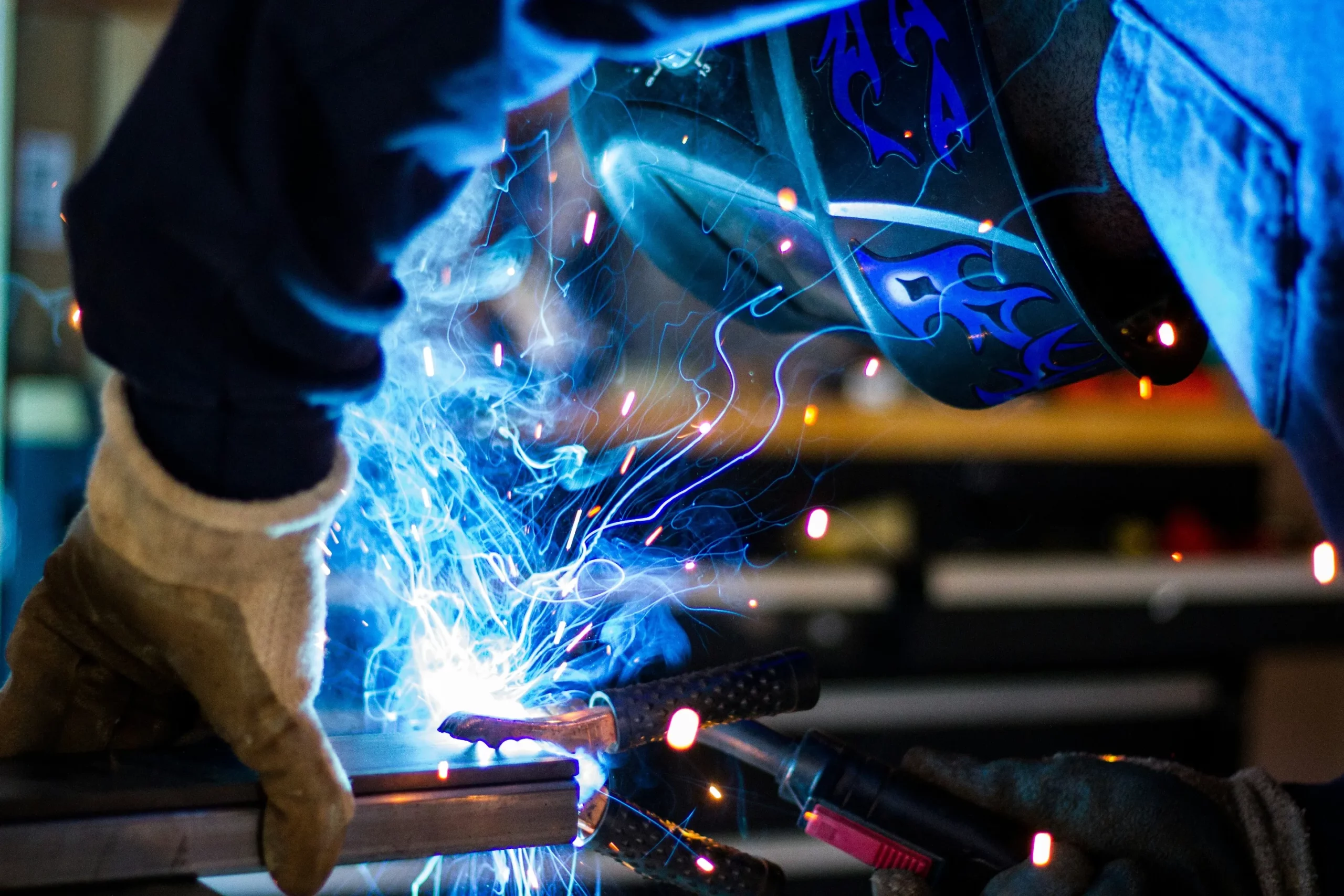A welder wearing protective gear, including gloves and a welding helmet with blue flame designs, uses a torch to weld a piece of metal. Bright sparks and blue light emanate from the welding process, creating a dynamic and intense scene.